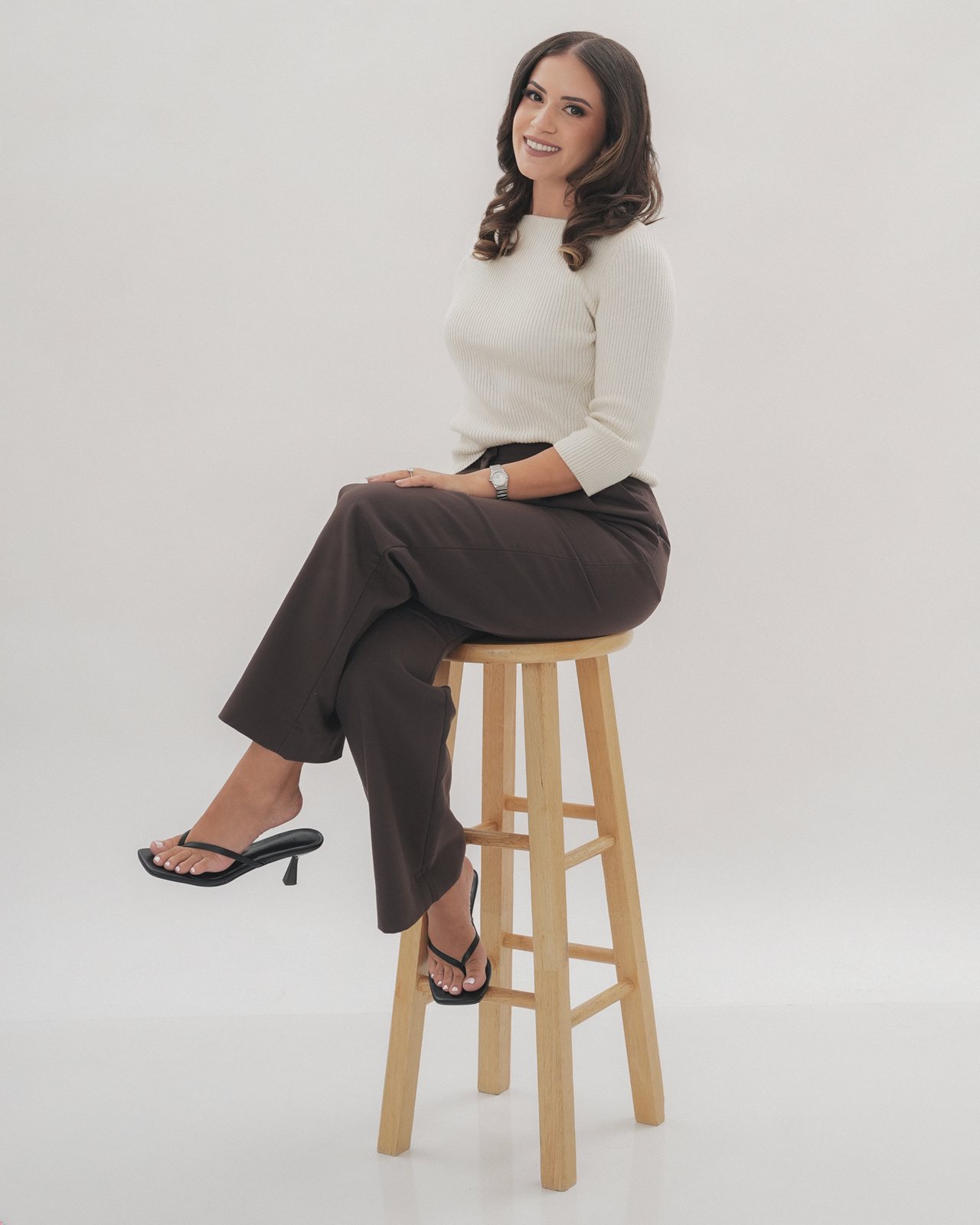 Abir Ayesh, LMFT, seated on a wooden stool with her legs crossed, smiling toward the camera.